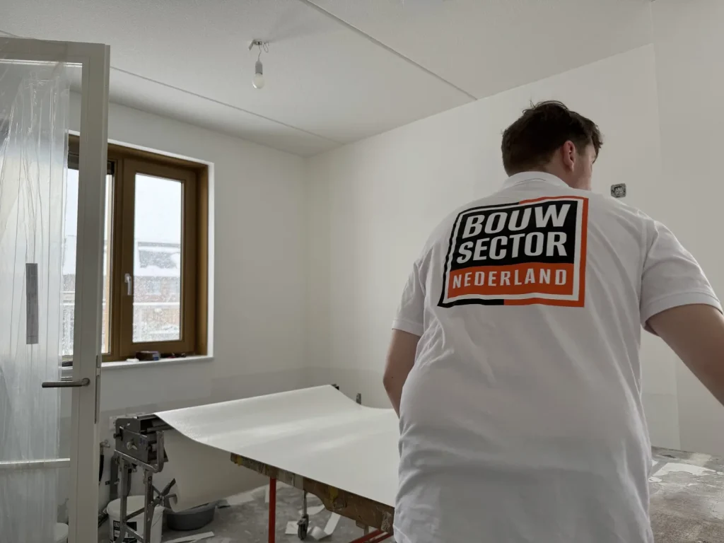 Behanger in een wit poloshirt met logo van Bouwsector Nederland rolt een baan vlies uit op de plaktafel