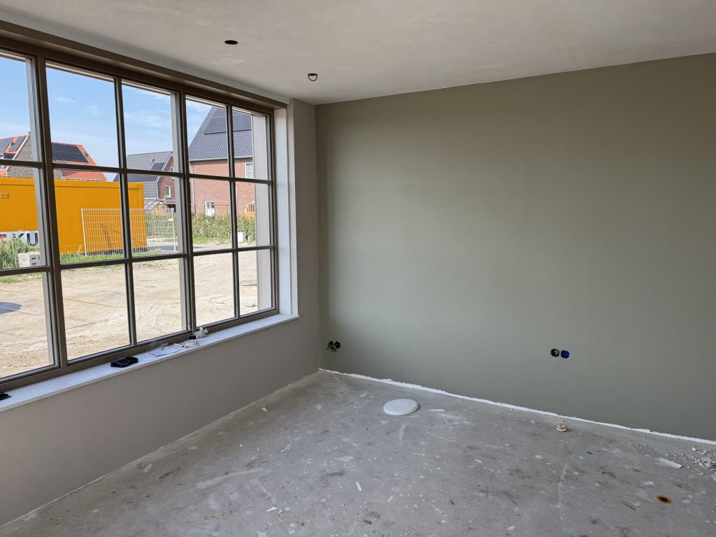 Nieuwbouwkamer met een egaal afgewerkte grijsgroene wand en grote ramen met uitzicht op nieuwe woningen