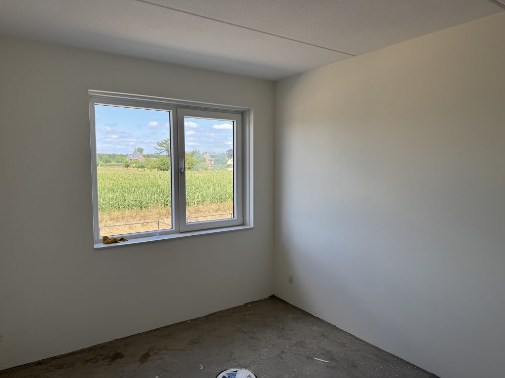 Lege nieuwbouwkamer met strakke lichte wanden en een raam met uitzicht op een maisveld
