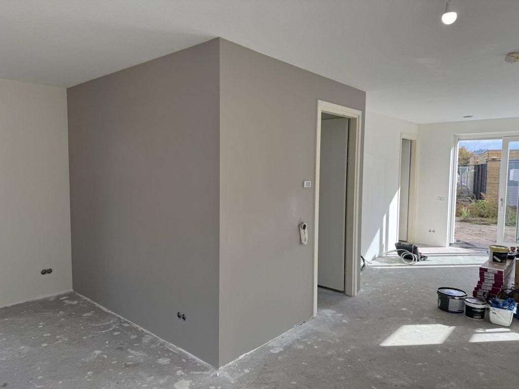 Open woonkamer in een nieuwbouwhuis met een pas afgewerkte grijze wand, een betonvloer en verfemmers bij de tuindeur